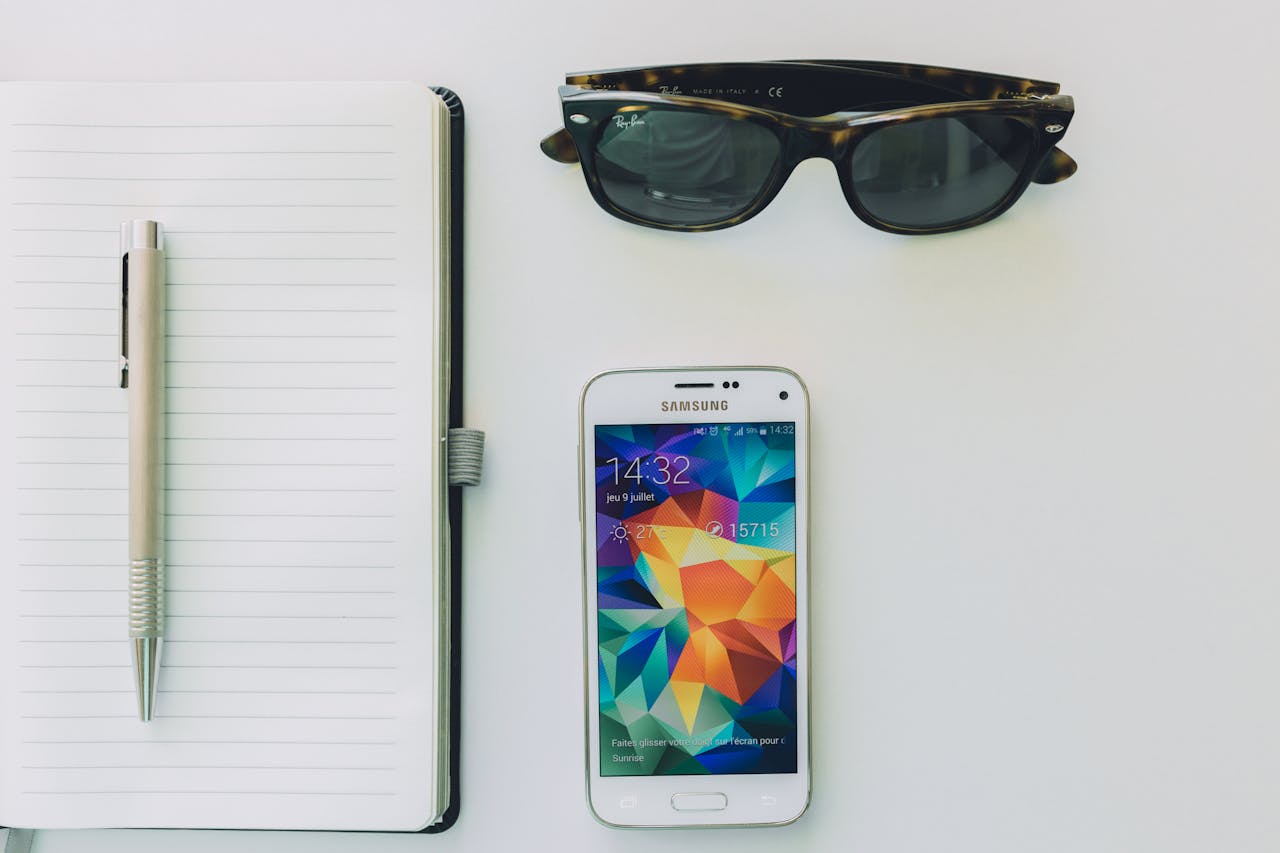 Flat lay image featuring a smartphone, notebook, and sunglasses on a white desk. Ideal for tech and productivity themes.